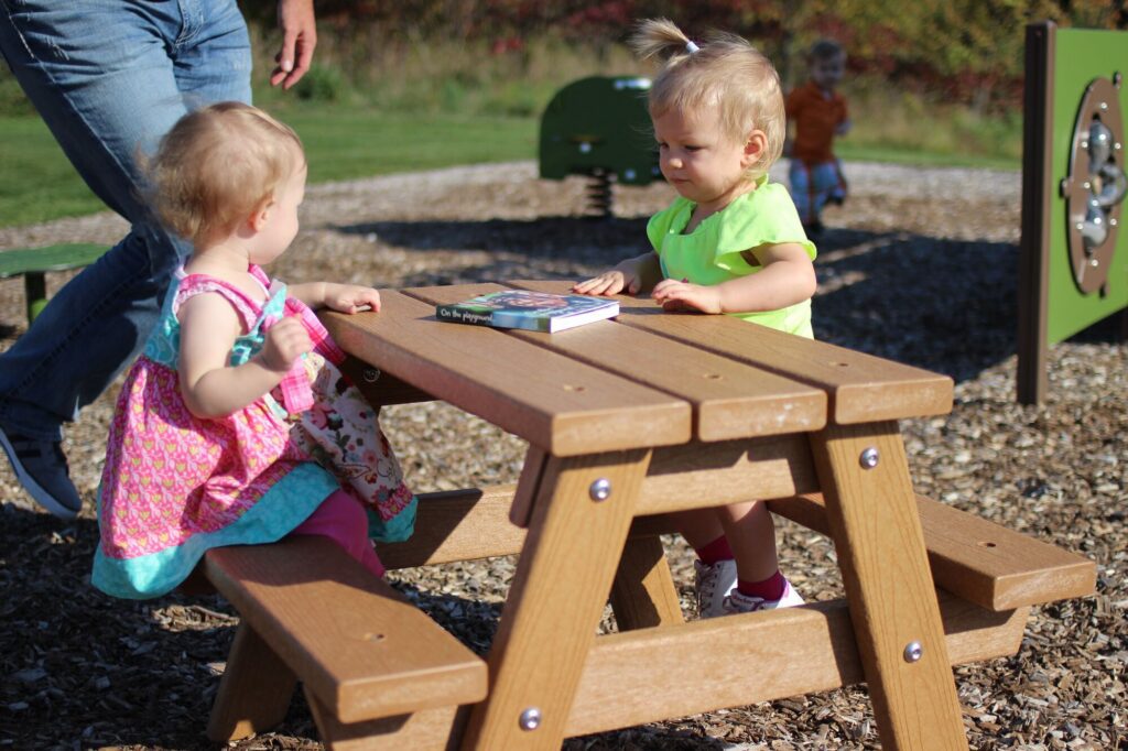 Two toddlers sit at an outdoor wooden picnic table with a book, while adults stand close by.