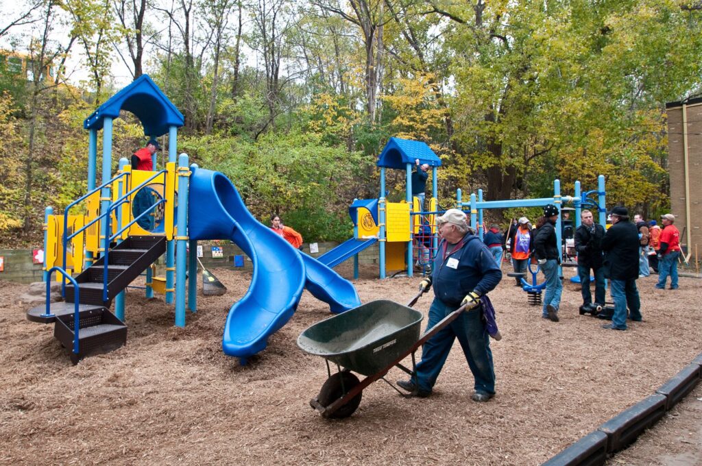 Volunteers build a playground with blue slides and yellow railings as children play in the background.
