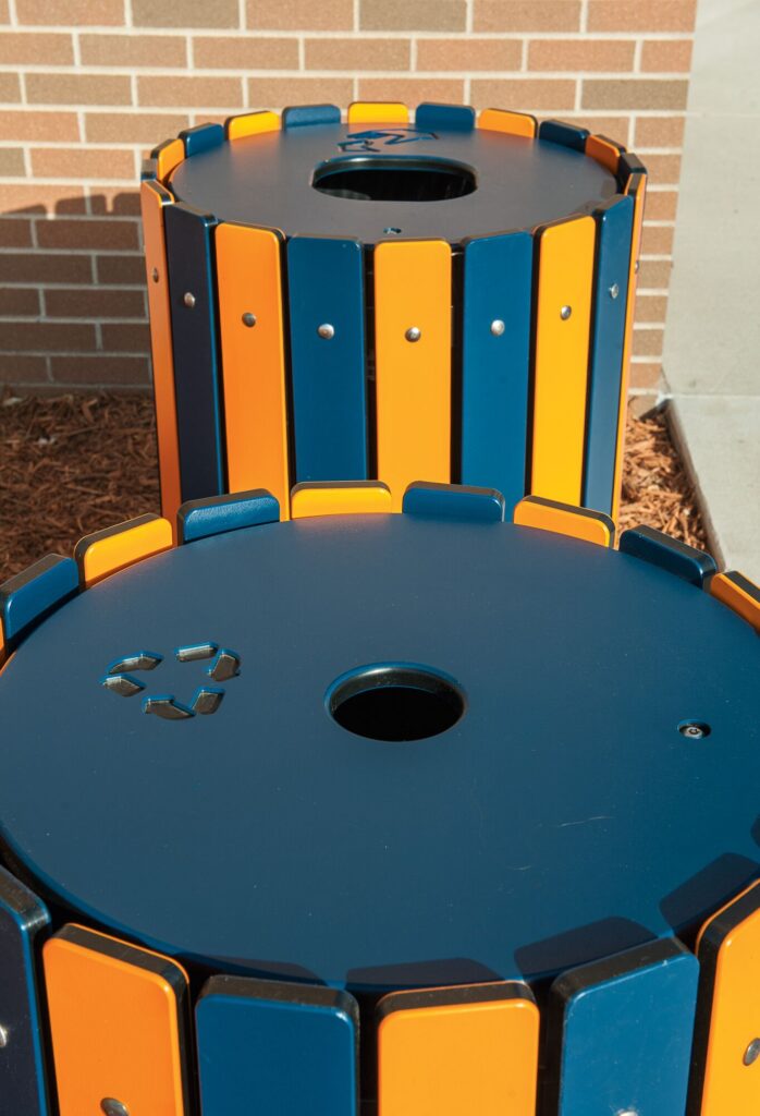 Blue and orange trash bins, one labeled for recycling, stand outdoors beside a brick wall.