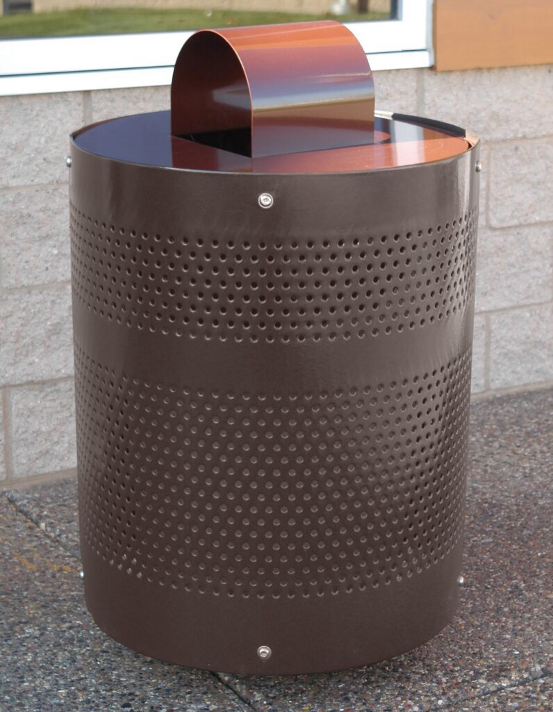 Brown metal trash can with perforated sides sits on a sidewalk next to a building wall.