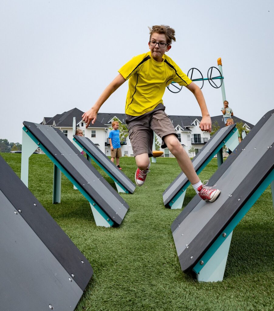 Boy in a yellow shirt jumps over angled platforms on a playground obstacle course as other kids play in the background.