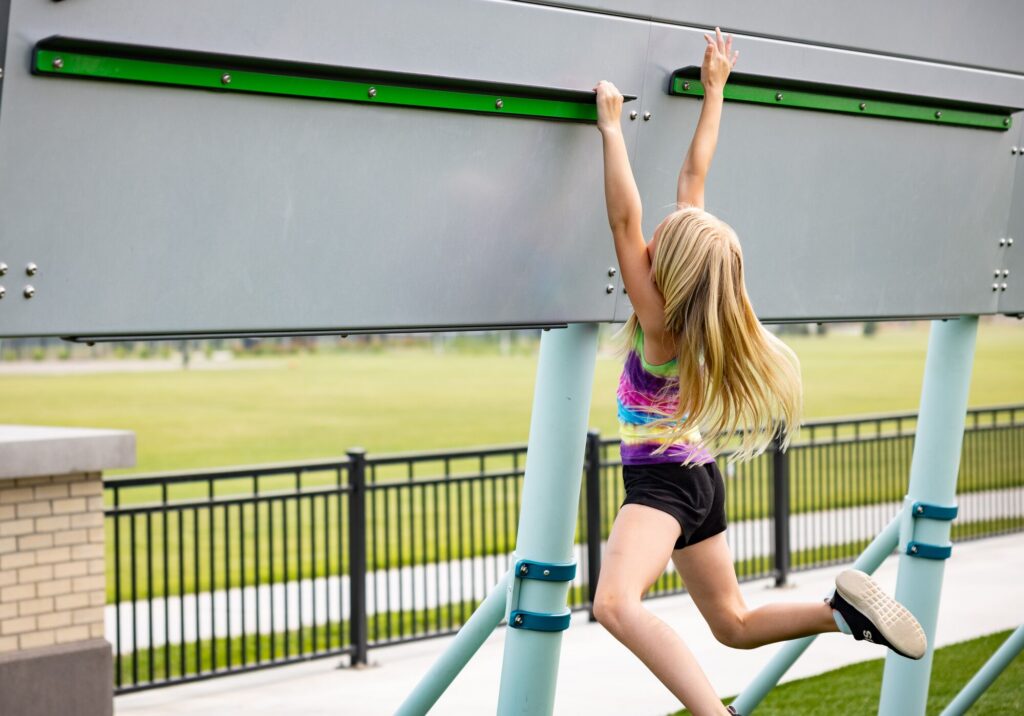 A young girl dangles from a playground climbing wall, reaching up with both hands as one foot hovers above the ground.