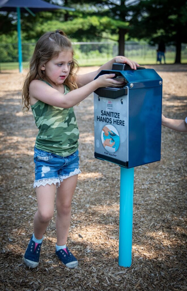 Girl in a park uses a hand sanitizer dispenser with a sign that reads Sanitize Hands Here.