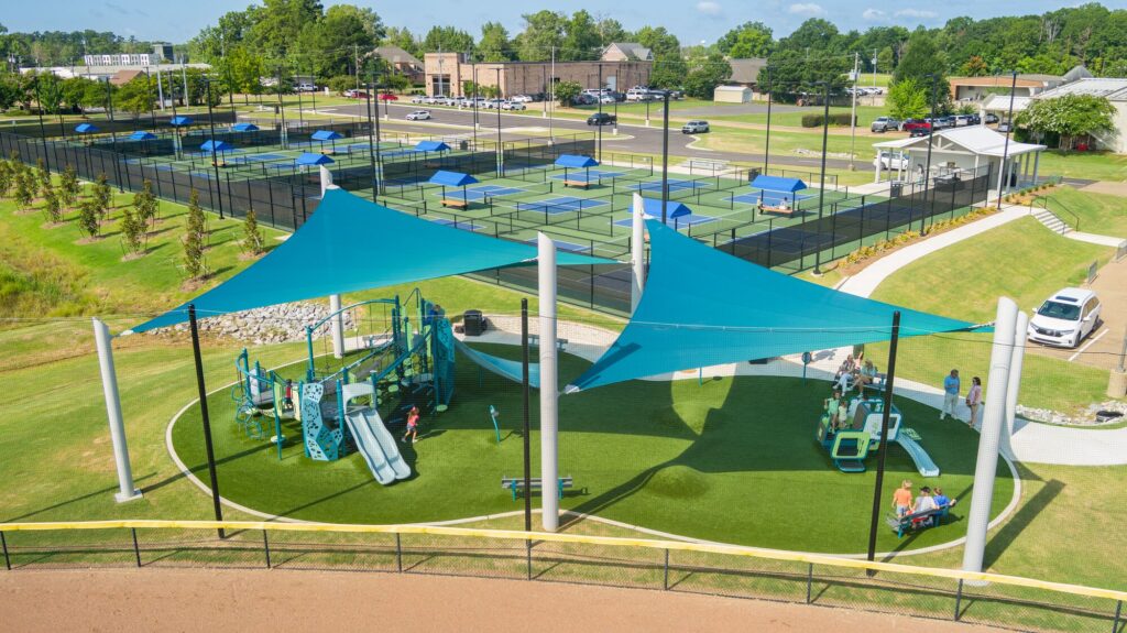 Blue shade sail playground beside tennis courts and parking lot, all under a clear sunny sky.