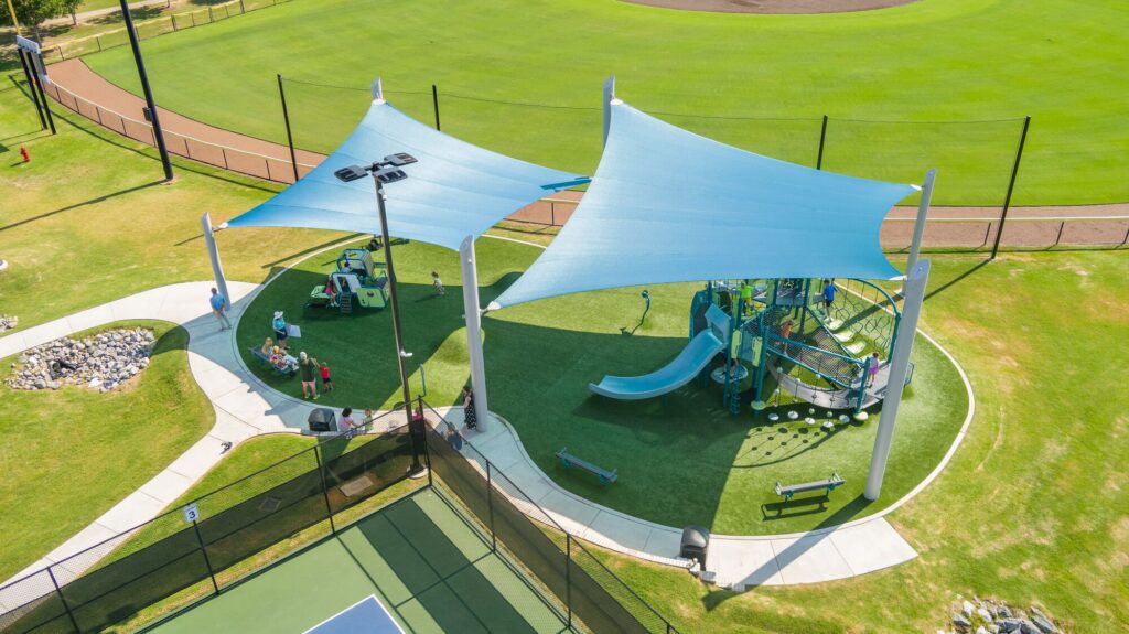 Aerial view of a playground with blue shade sails, slides, and kids playing on bright green artificial turf.