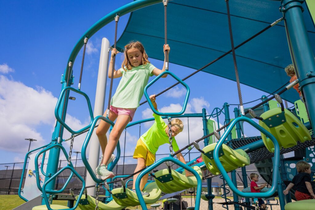 Children climb a geometric jungle gym at an outdoor playground beneath a blue canopy on a sunny day.