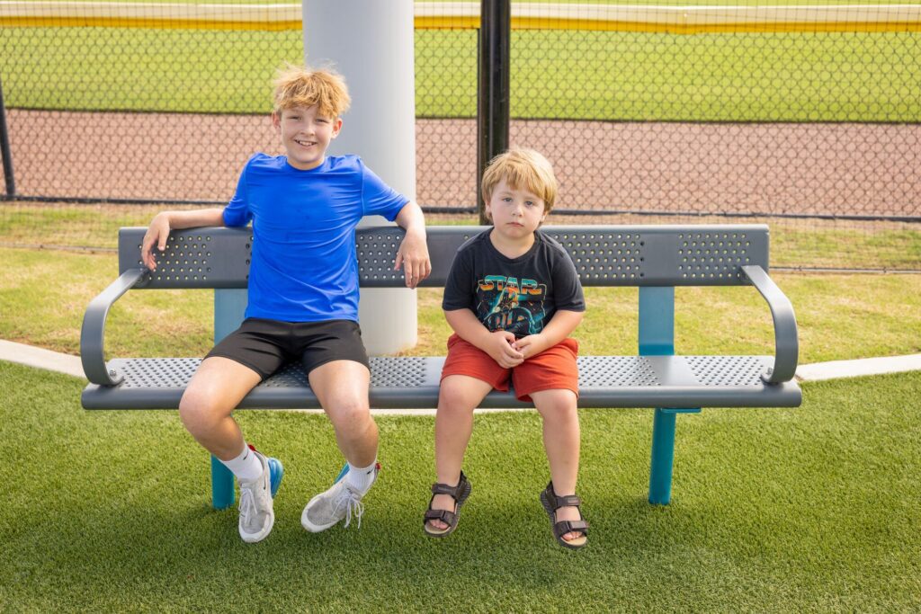 Two boys sit on a park bench by a baseball field; one smiles while the other has a serious expression.