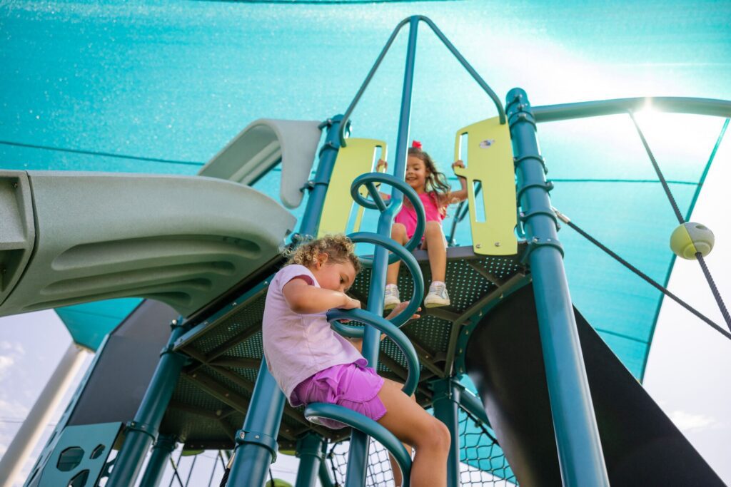Two young girls climb and play on a playground structure shaded by a blue canopy on a bright, sunny day.
