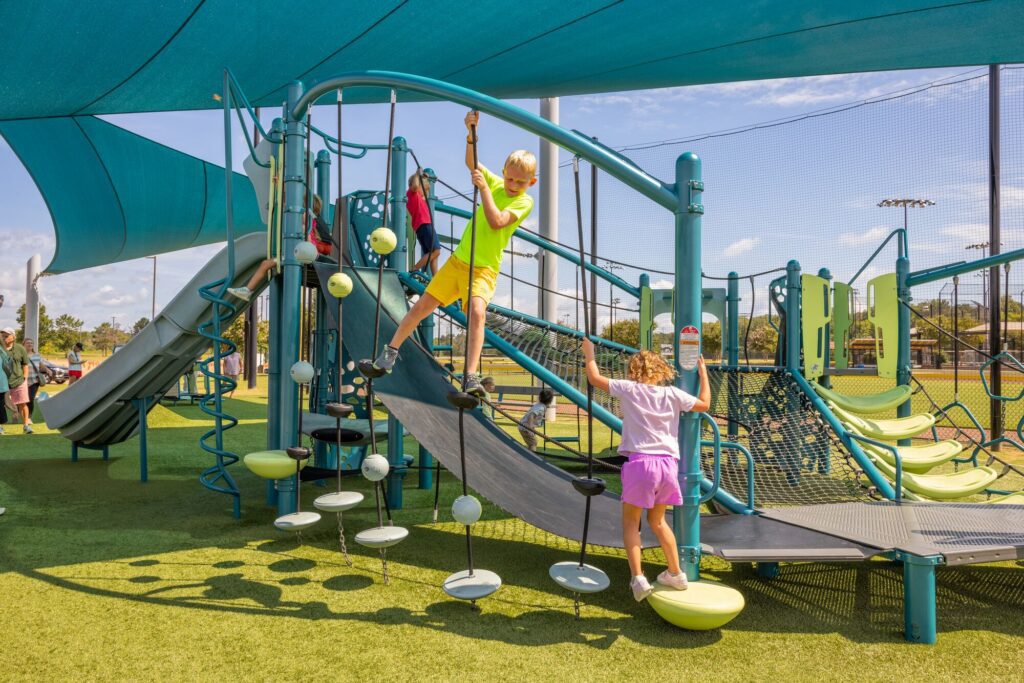 Two kids play on a modern playground under blue shade sails, sunlight casting bright patches on the ground.