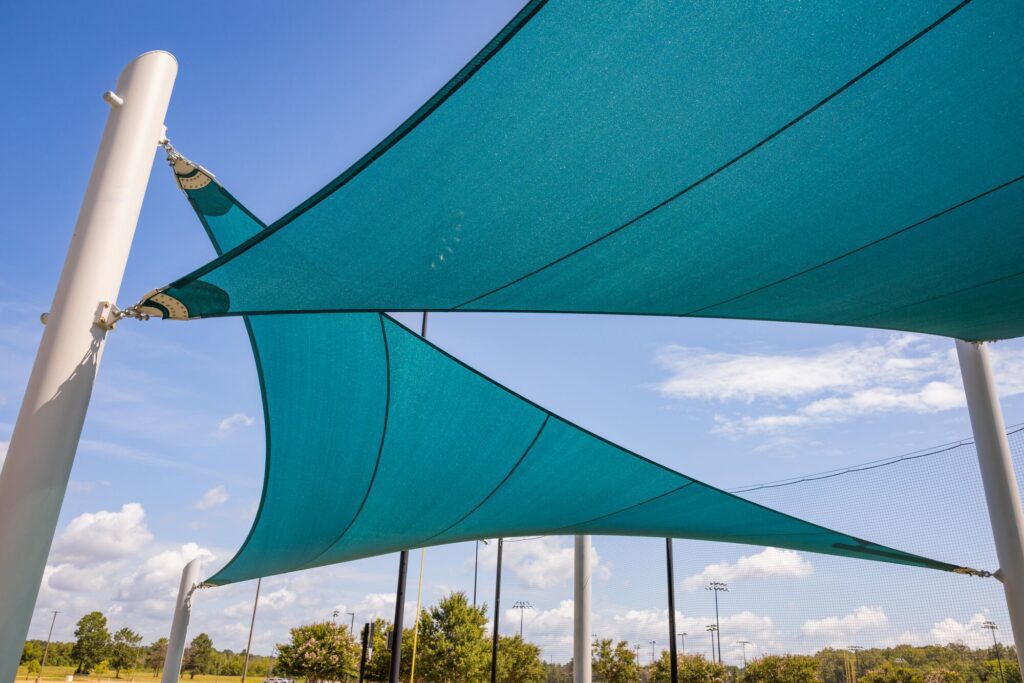 Blue shade sails span white poles beneath a sunny sky, with trees and scattered clouds in the background.
