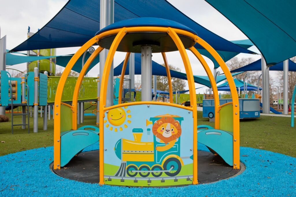 Bright playground with a lion driving a train, blue shade sails above, and green artificial turf underneath.