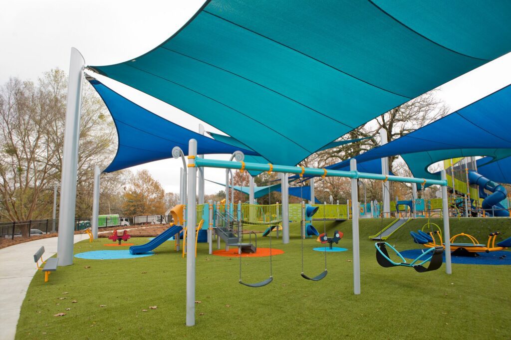 Playground featuring swings, slides, climbing frames, and blue shade sails above artificial turf.