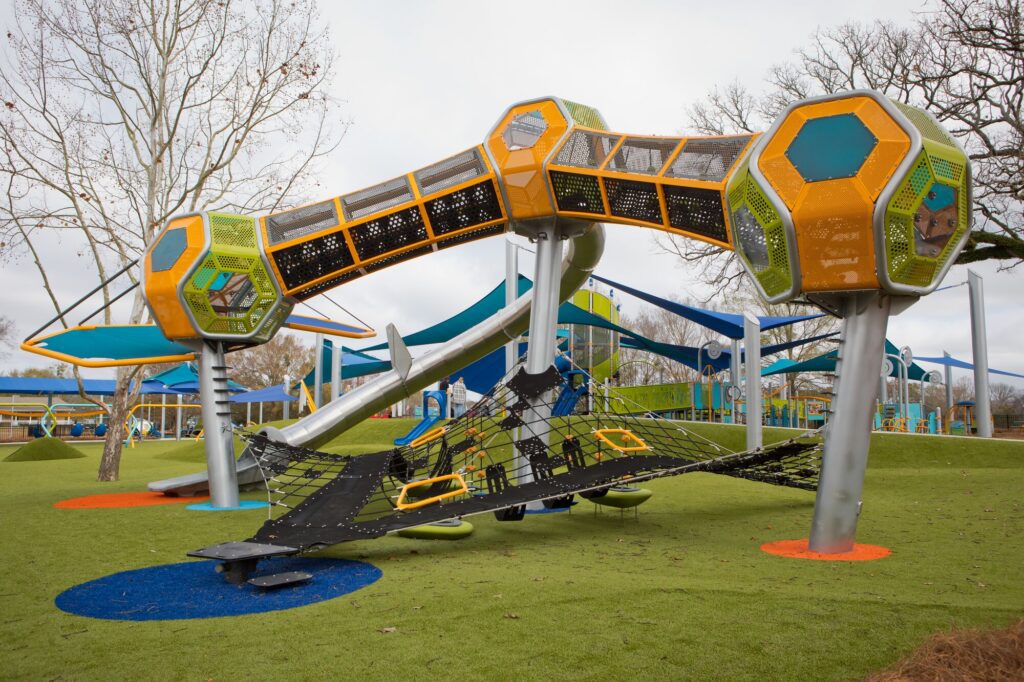 Modern playground features bright climbing nets, slides, and geometric play structures on green artificial turf.