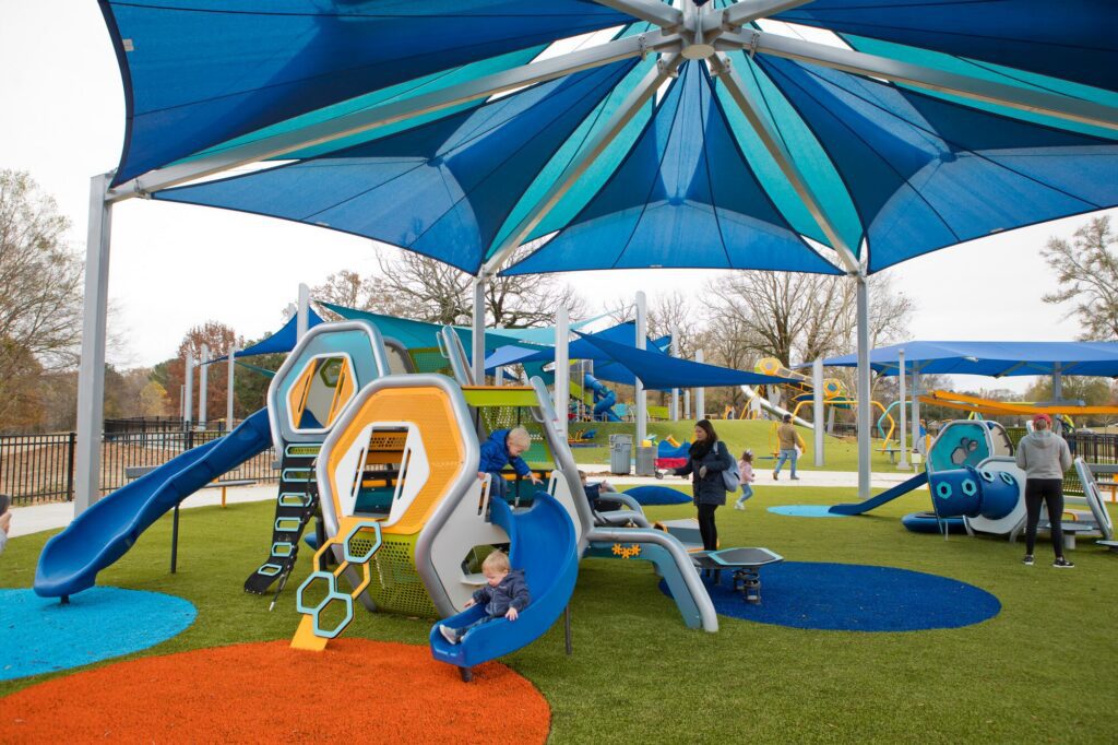 Kids play on bright, modern playground structures shaded by big blue canopies in a sunny park.