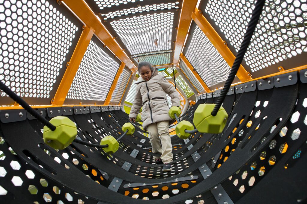 Child in a coat climbs through a modern playground tunnel with yellow and green geometric shapes.
