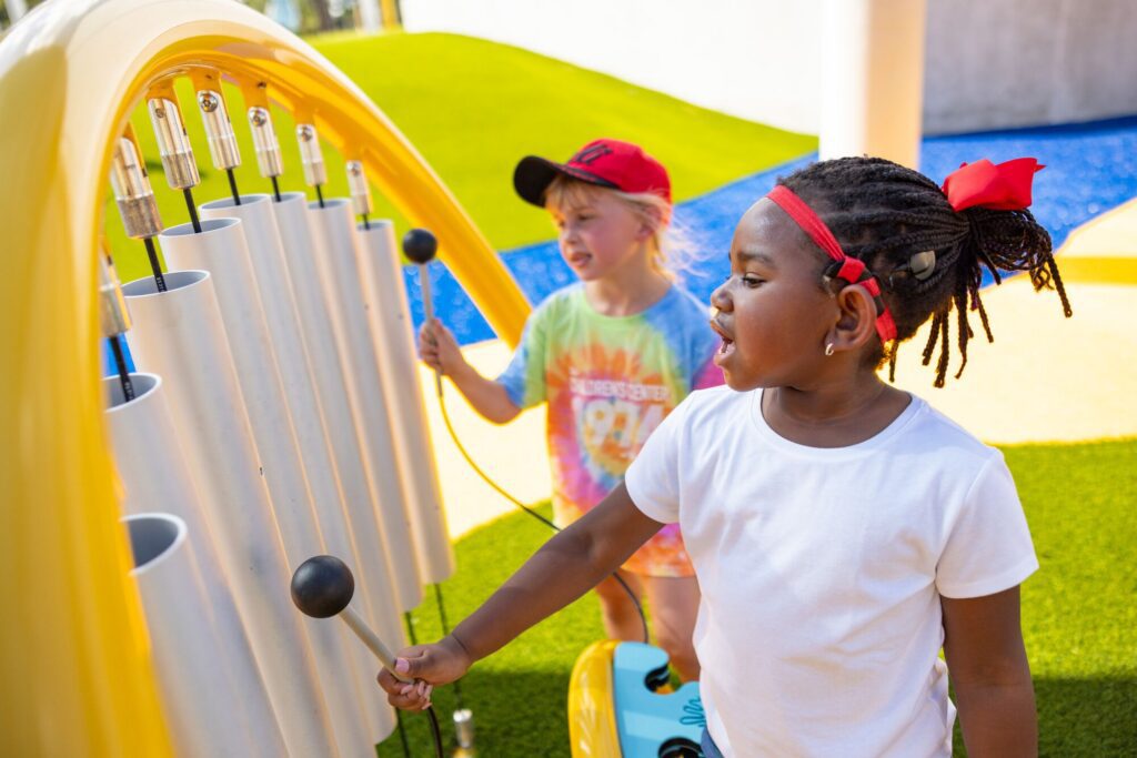 Two kids at a playground play outdoor chimes; one strikes the tubes while the other watches.