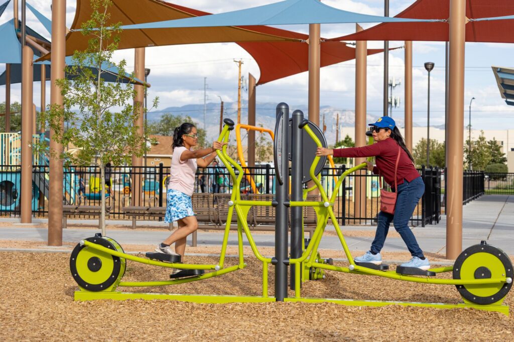 Two people use a green outdoor exercise machine at a playground beneath bright, colorful shade canopies.