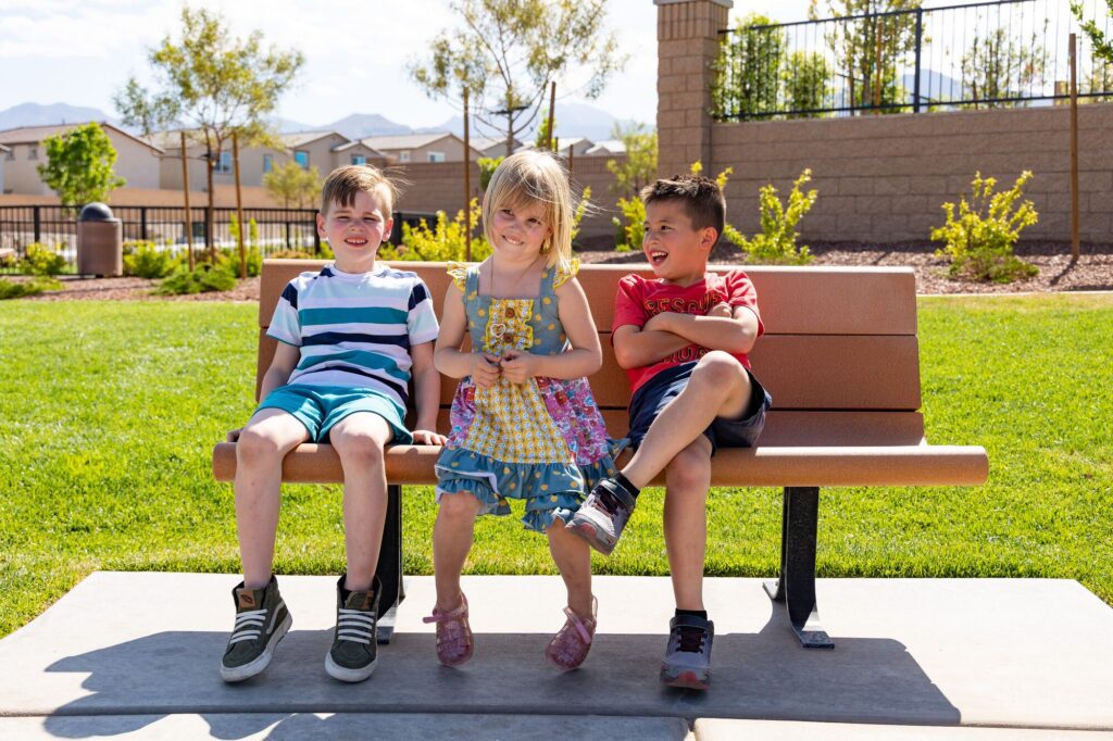 Three children smile as they sit together on a park bench, enjoying a sunny day outdoors.