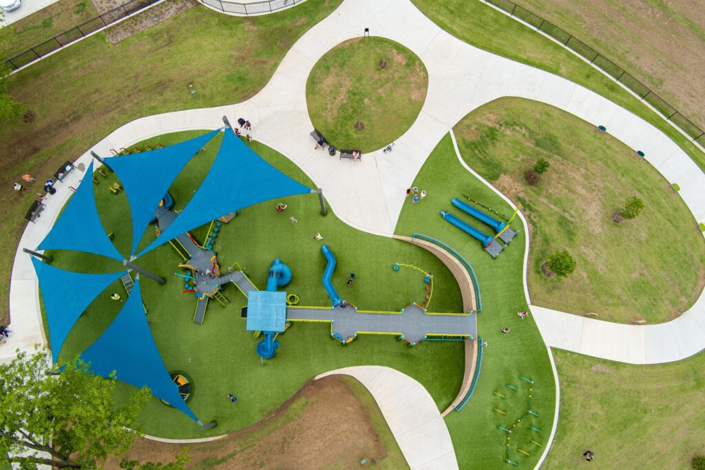 Aerial view of playground featuring blue shade sails, green turf, and curved concrete walkways.