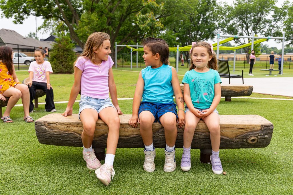 Three smiling children sit on a log bench in a sunny park, chatting together.