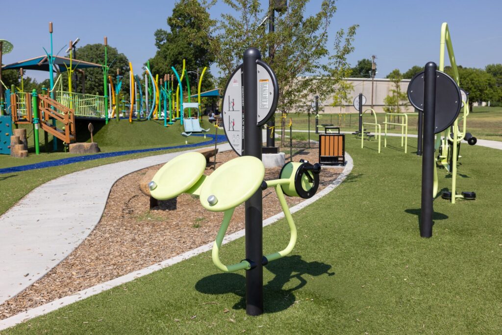 Outdoor gym equipment sits on artificial grass by a park path, with playground structures visible in the background.