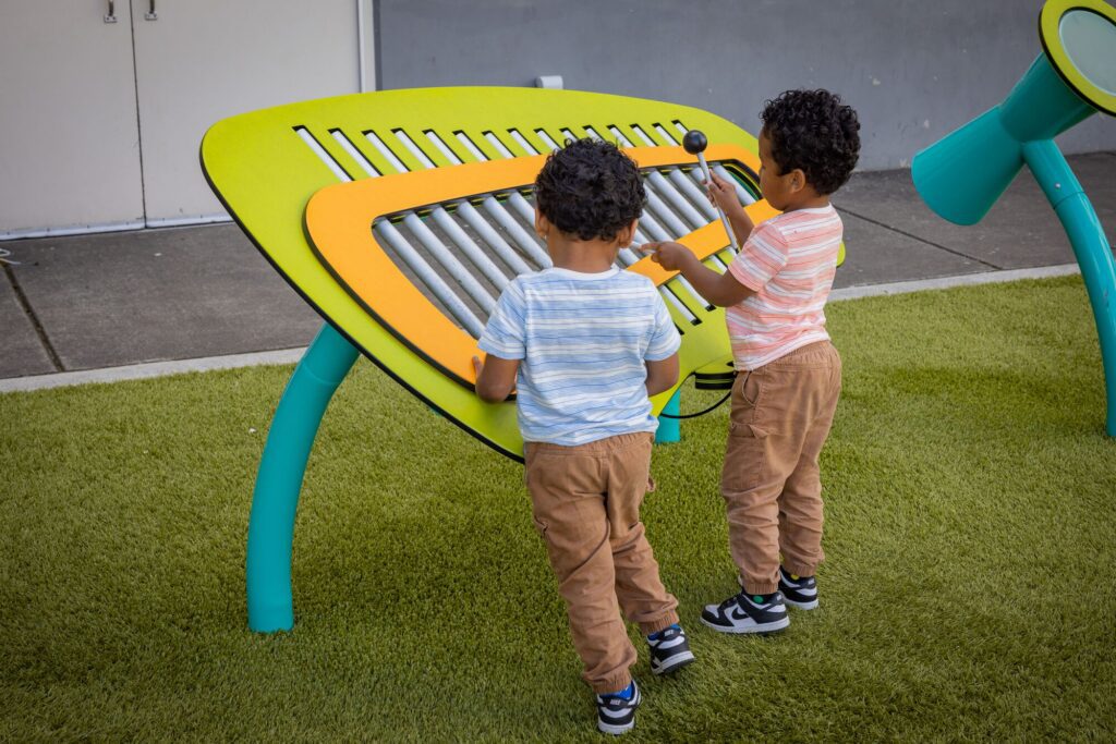 Two young kids play a big outdoor xylophone together on green artificial grass.