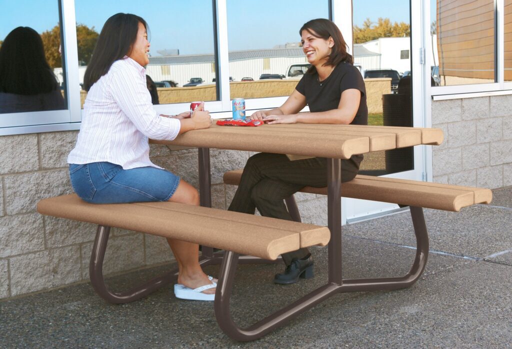 Two women chat and smile at an outdoor picnic table, drinks and snacks scattered in front of them.