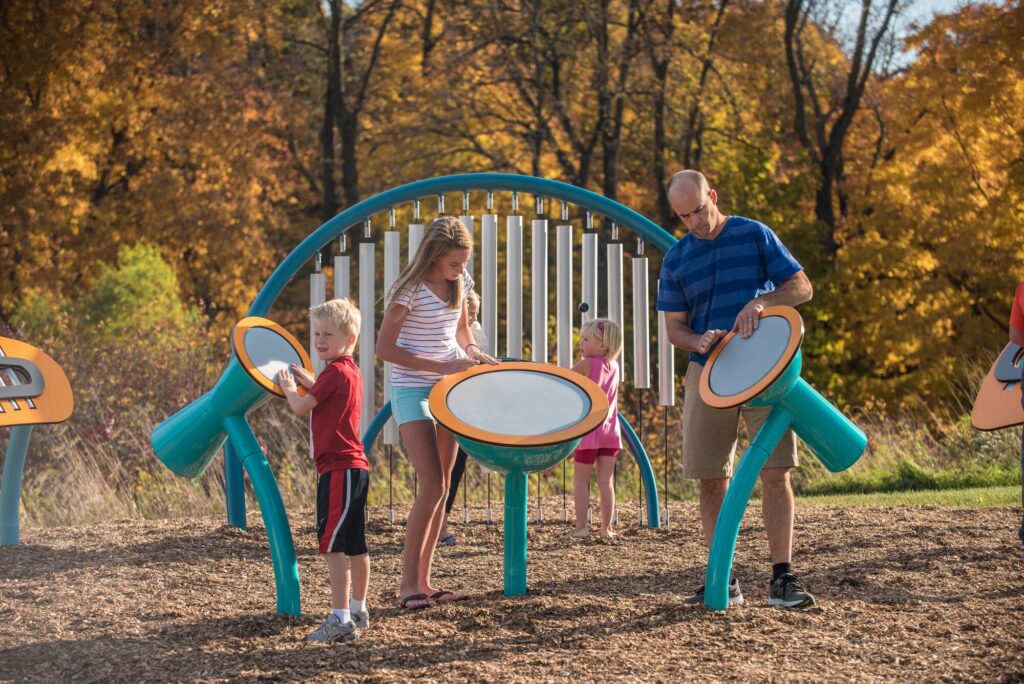 Kids and adults play drums at an outdoor playground, framed by colorful autumn trees.