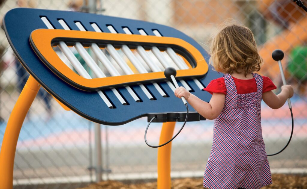 A small child uses mallets to play a colorful, oversized xylophone at a playground.