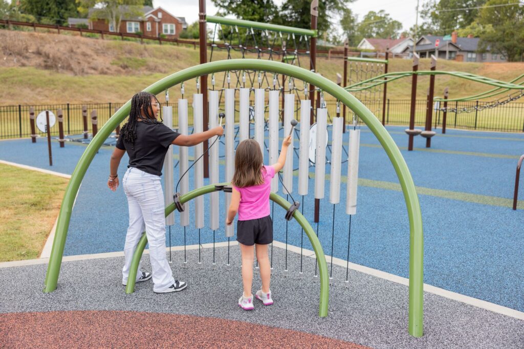 Two girls play a big chime instrument on a playground with blue rubber flooring.