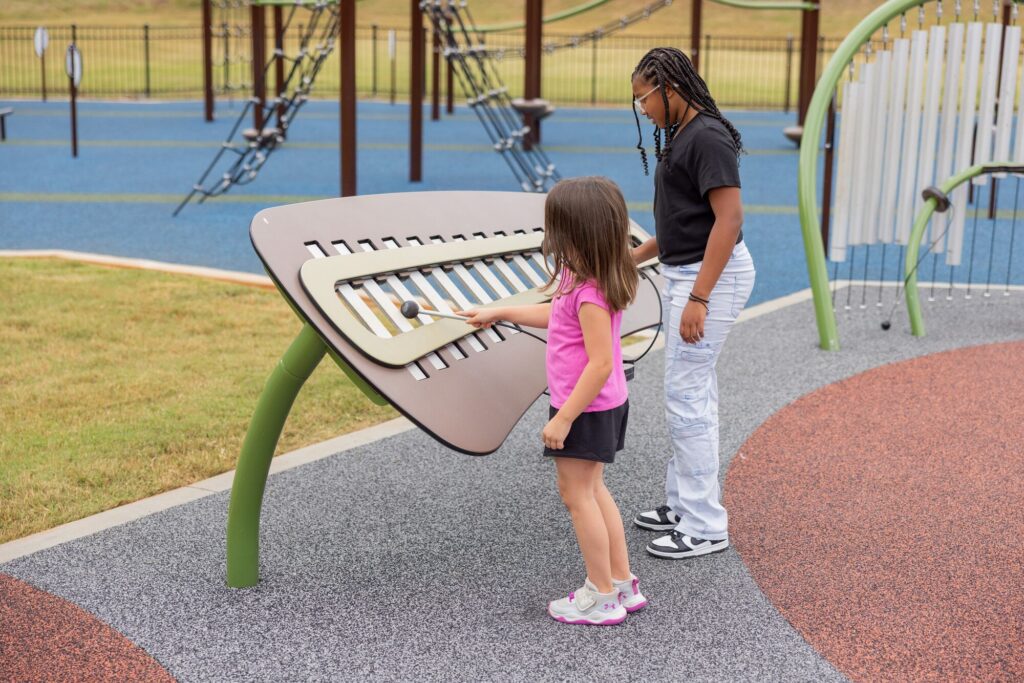 Two kids play a big outdoor xylophone on a rubber playground surface.