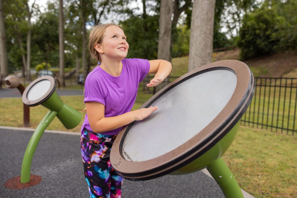 Smiling girl in a purple shirt plays a big outdoor drum at a playground, trees visible in the background.