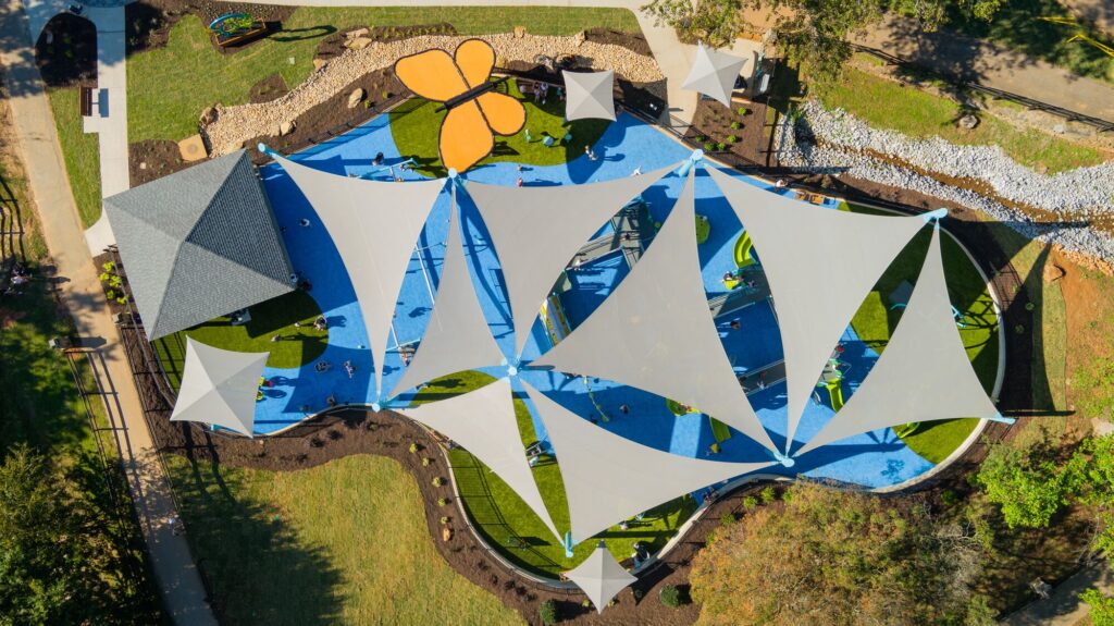 Playground with blue floor and white shade sails, seen from above, surrounded by lush green trees.