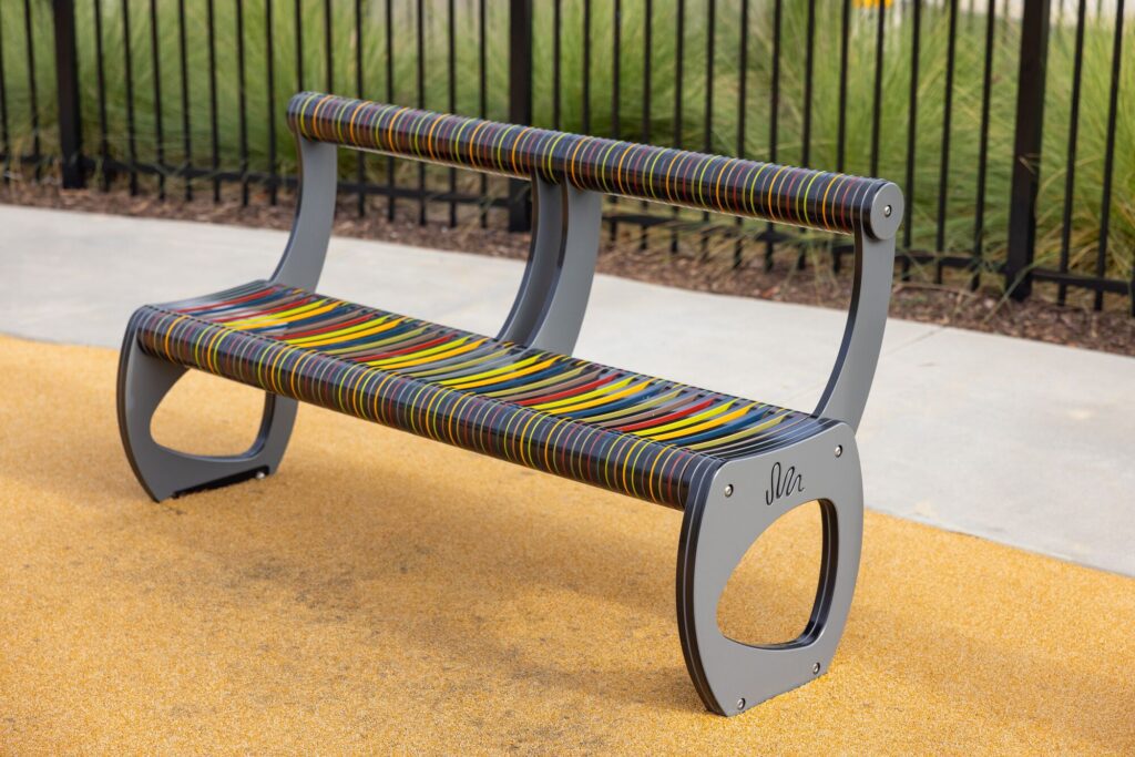 Modern bench with multicolored striped slats sits on yellow ground by a black fence and green grass.