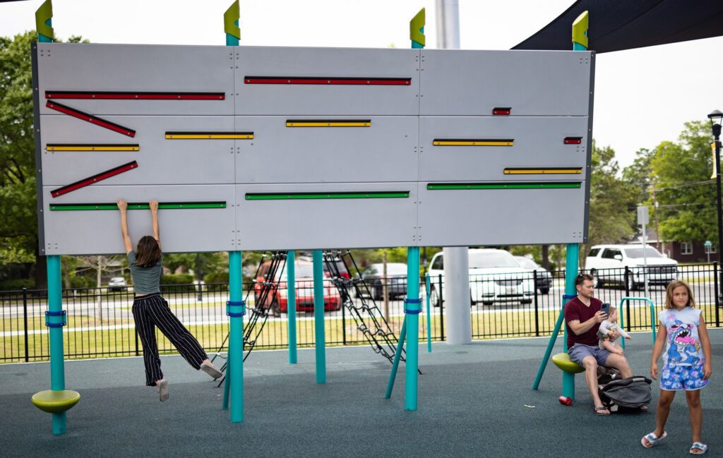 Child swings from big playground wall while other kids play and sit nearby on a bright, sunny day.