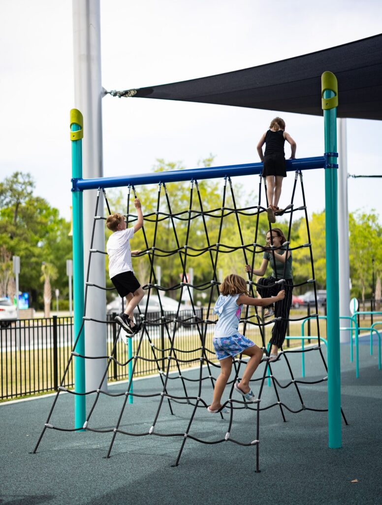 Four kids scale a rope net at an outdoor playground under bright sunlight.