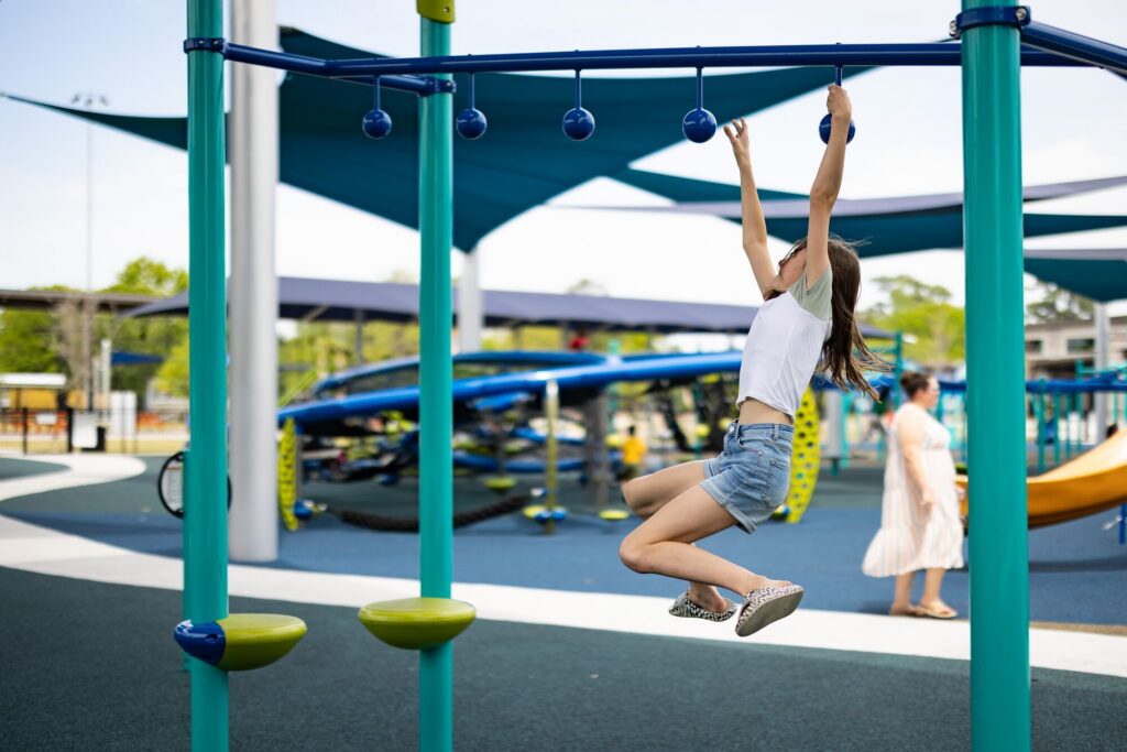 Girl in shorts and t-shirt hangs from monkey bars at a playground, play structures visible behind her.