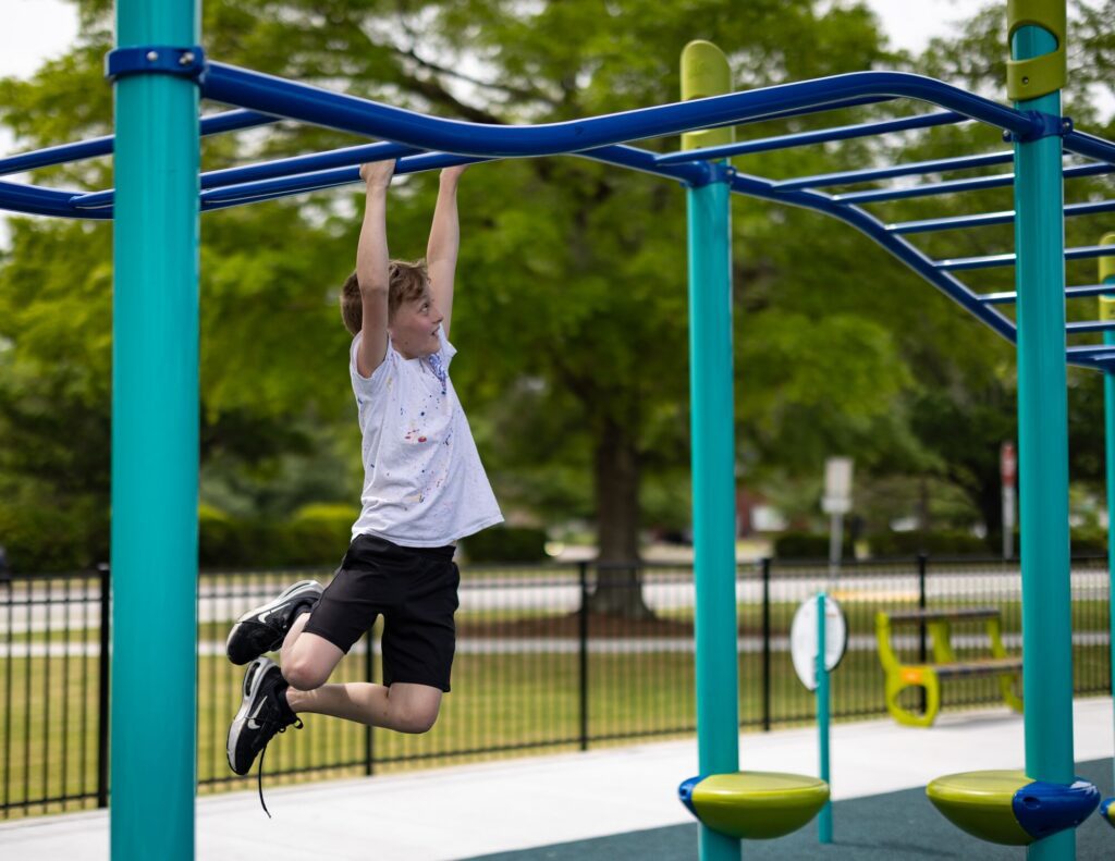 Boy wearing a white shirt and black shorts hangs from monkey bars at a playground, green trees in the background.
