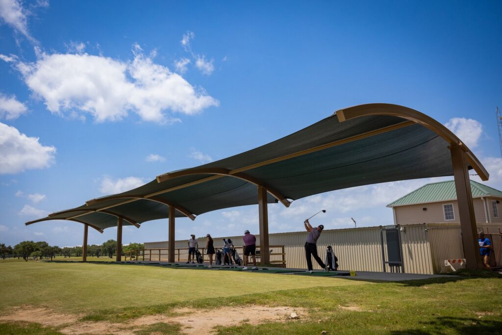 Golfers swing under a big shaded canopy at a driving range on a bright, sunny day.