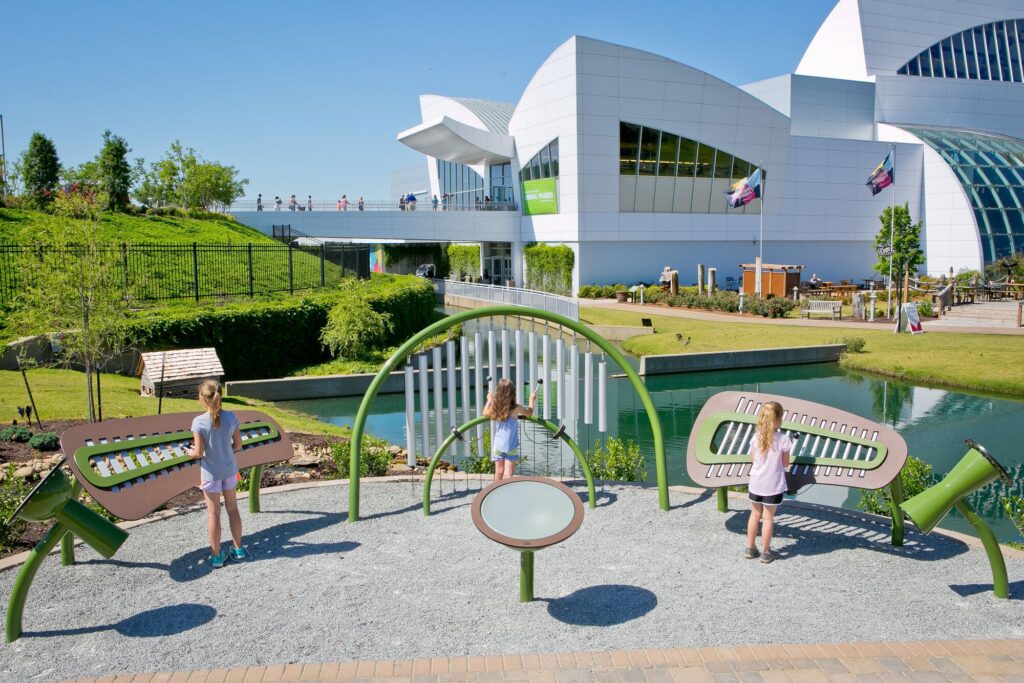 Children play outdoor musical instruments by a pond, with a modern white building in the background.