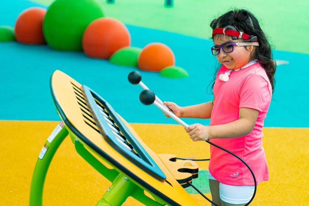 Girl in glasses uses mallets to play a large outdoor musical instrument on a brightly colored playground.