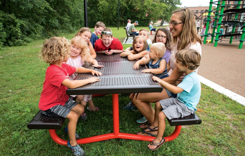 Children and an adult woman smile and laugh together around a picnic table outdoors in a park.