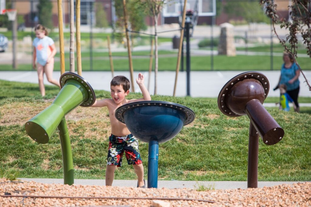 Shirtless boy in swim trunks plays on park equipment while other children are visible in the background.
