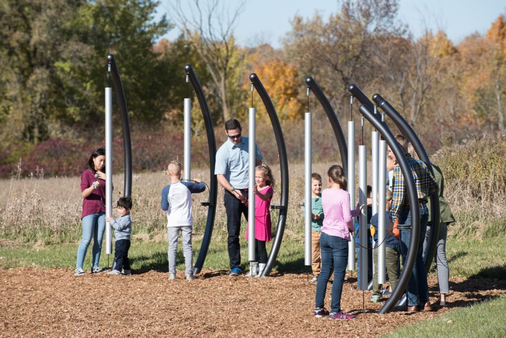 Kids and adults play on a musical playground in a sunny park, enjoying instruments together.