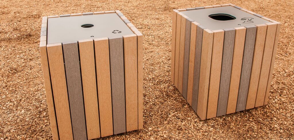 Two wooden recycling and waste bins sit side by side on a mulch-covered ground outdoors.