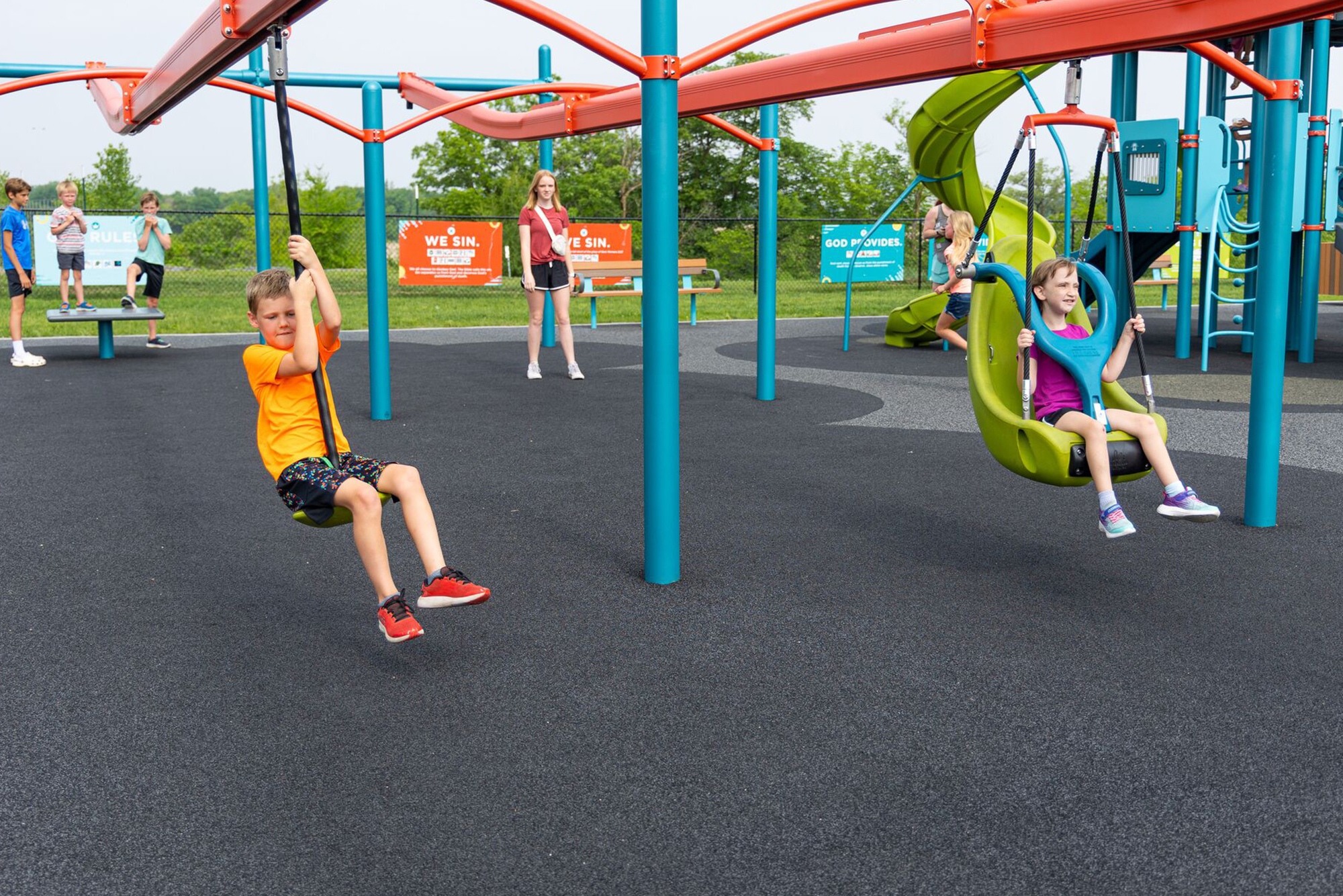 Two kids play on swings in a bright playground as other children and adults watch in the background.