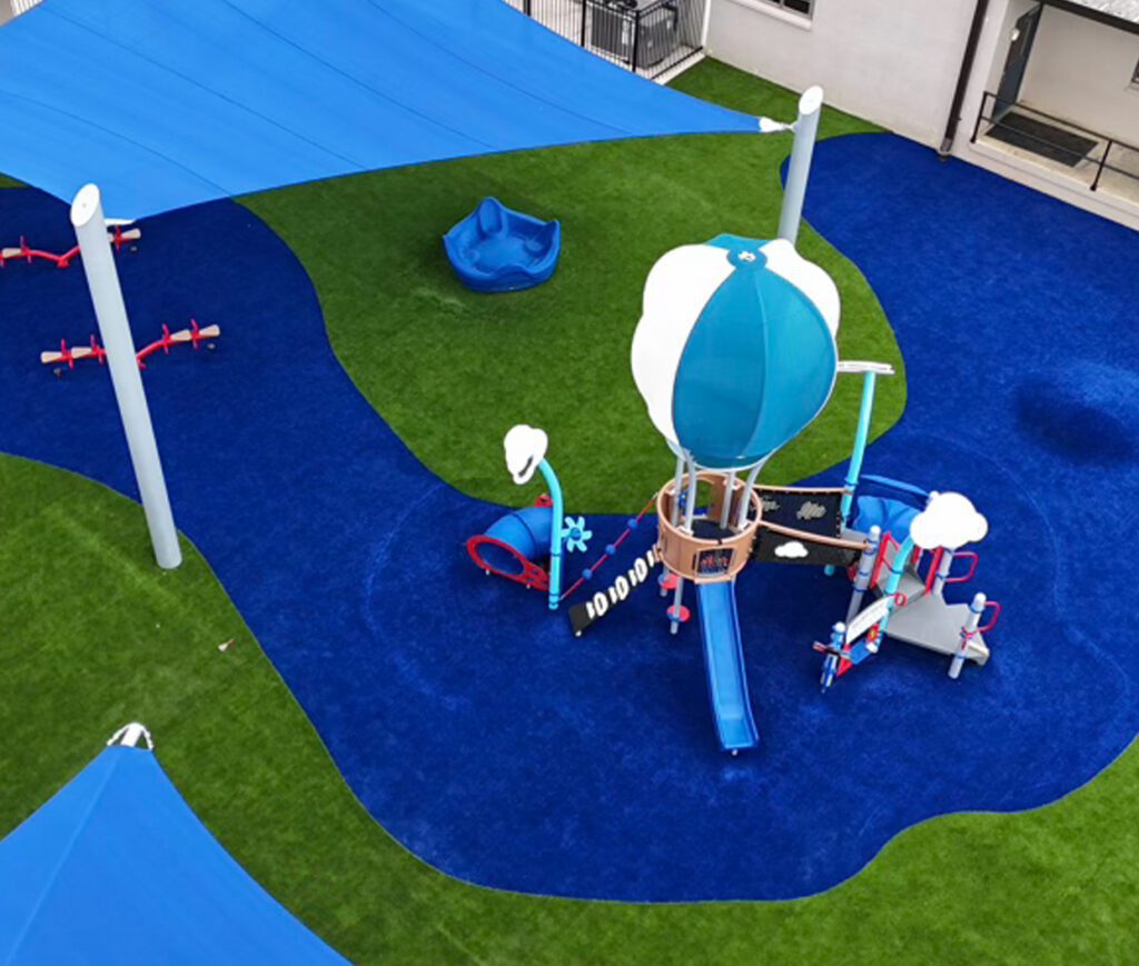 Aerial view of a playground with blue turf, slides, seesaws, and shade canopies on synthetic grass.
