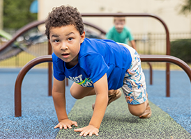 A young boy crawls beneath playground bars, while another child plays in the background.