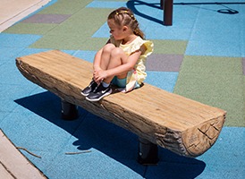 Young girl in a yellow dress sits on a log bench at a playground with blue and green rubber flooring.