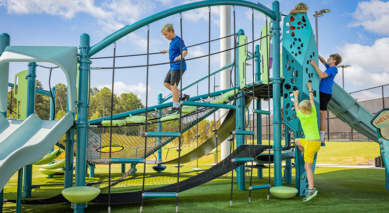 Four kids play together on a modern playground with slides, climbing walls, and nets under the bright sun.